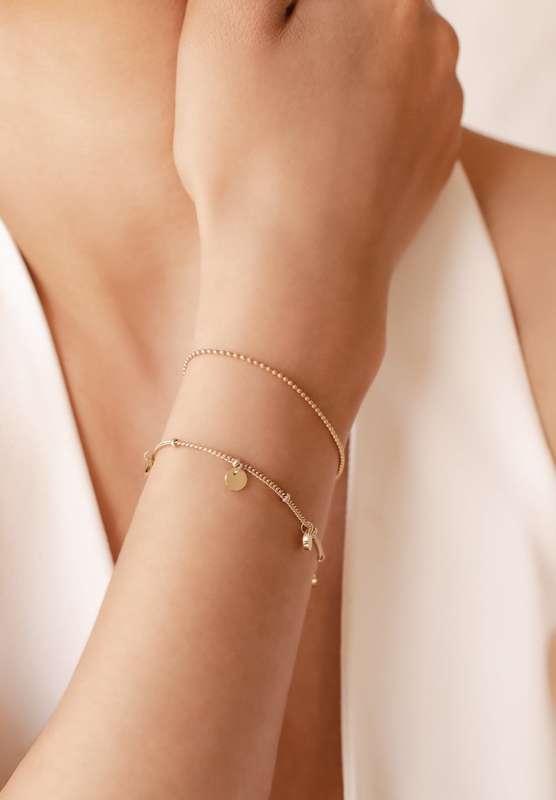 Goldfarbene Armbänder, eines mit kleinen Perlen, das andere mit einem glatten Scheiben-Anhänger und zarter Kette, vor einem hellen Hautton.
