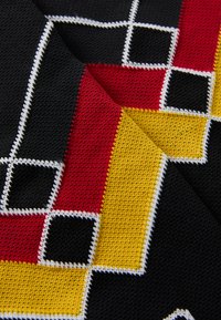 Close-up de tecido tricotado com padrão geométrico em preto, vermelho, amarelo e branco, apresentando quadrados e linhas diagonais.