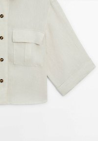 Short-sleeve, light beige shirt in a textured fabric, featuring a button-down front and a single chest pocket. Brown buttons add contrast.