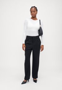 Black tailored trousers with a high waist, pleated front, and sturdy fabric. Paired with a fitted white long-sleeve top and black handbag.