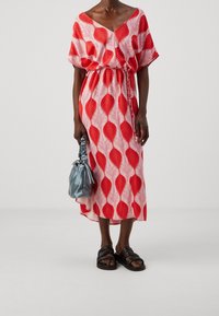 Robe à motifs de feuilles rouges et blanches avec une coupe fluide et une taille ceinturée, associée à un sac bleu clair et des sandales plates noires.