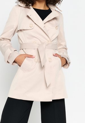 Vrouw draagt een beige dubbelgeknoopte trenchcoat met ceintuur en zwarte broek, handen in de zakken van de jas tegen een witte achtergrond.