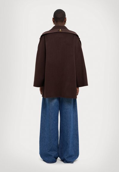 Hnědý vlněný kabát s širokými rukávy, s límcem a trojúhelníkovým akcentem na zadní straně. V kombinaci s objemnými modrými denimovými kalhotami.