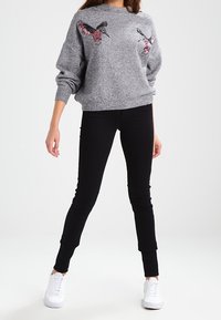 Modèle portant un pull gris avec des motifs d'oiseaux en sequins, associé à un jean slim noir et des baskets blanches, se tenant devant un fond uni.