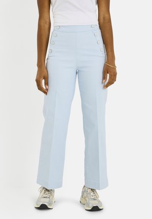 Mujer que lleva pantalones azul claro de talle alto con seis botones blancos en los lados delanteros, combinados con zapatillas blancas y una camiseta blanca.