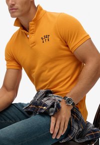 Oranje poloshirt met een kraag, voorzien van zwart geborduurde tekst. Blauwe jeans met een grijs en marineblauw geruit overhemd om de taille geknoopt. Zwarte horloge.