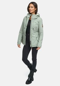 Light green hooded jacket with a zip-front, adjustable drawstrings, and two side pockets. Worn over a black top and paired with black jeans and boots.