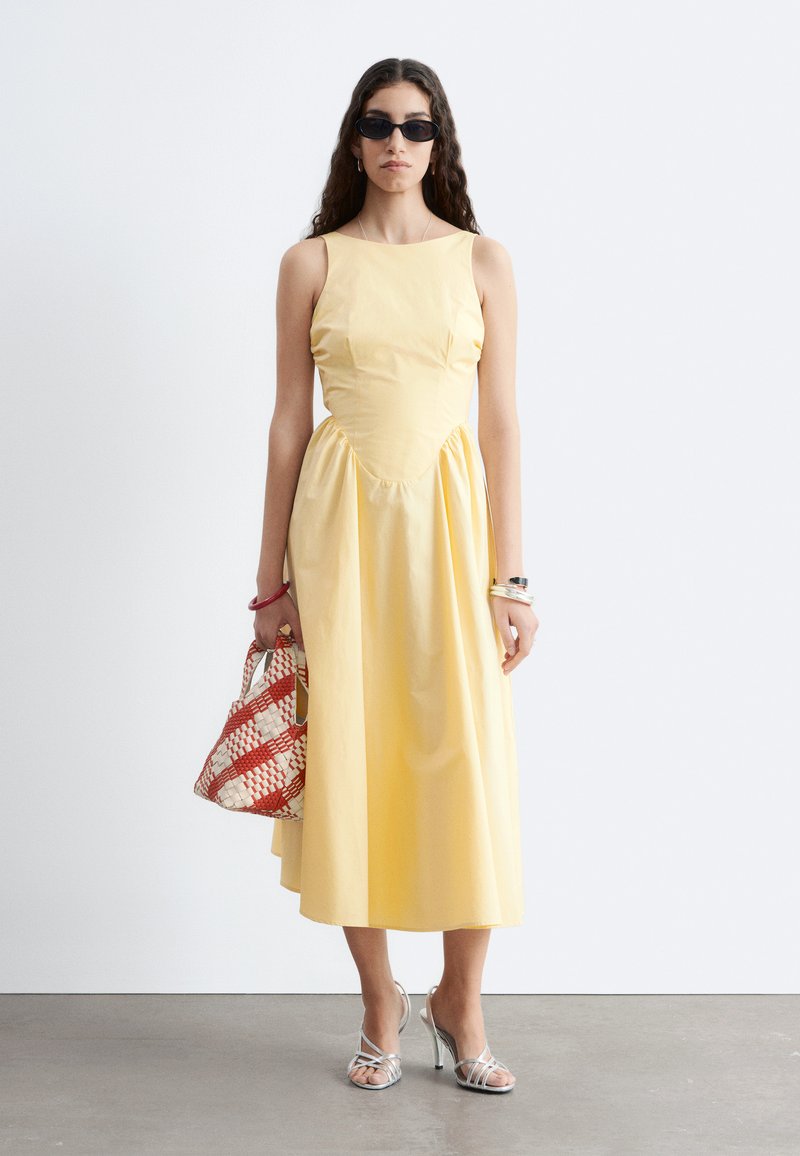 Femme portant une robe midi jaune sans manches, des lunettes de soleil noires, des sandales argentées à talons hauts et tenant un sac à main tissé rouge et blanc.