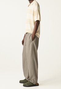 Korte mouw polo shirt met beige en crème strepen, gecombineerd met losse lichtgrijze broek en donkergroene klompen met een gespsluiting.