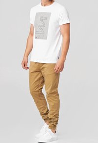 Weißes Baumwoll-T-Shirt mit schwarzen grafischen Linien, kombiniert mit hellbraunen Jogginghosen und weißen Sneakers, das ein lässiges Outfit zeigt.