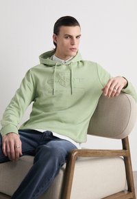 Sudadera verde claro con un logotipo frontal, textura suave, cordones y puños acanalados. Combinada con jeans oscuros, usada de manera informal sobre una silla.