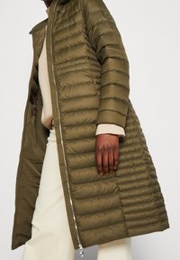 Persoon in een lange olijfgroene gewatteerde jas over een beige trui en witte broek, staande tegen een effen achtergrond.