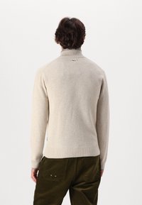 Beżowy sweter dzianinowy z wysokim kołnierzem i ściągaczem. Posiada subtelny detal w postaci czerwonego i niebieskiego logo na plecach. Teksturowany materiał oraz dopasowany krój.