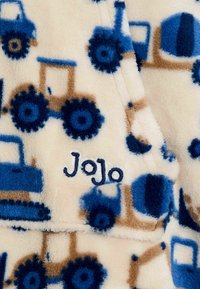 Fleece-stof med blå traktor- og lastbilmønstre på en cremefarvet baggrund, med broderet "Jojo" i blå tråd. Blød tekstur.
