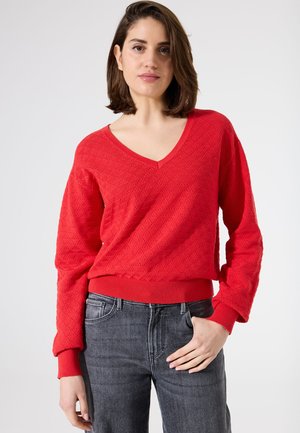 Rode gebreide trui met een V-hals, gestructureerd ruitpatroon en rib gebreide boorden en zoom, gecombineerd met donkergrijze jeans.