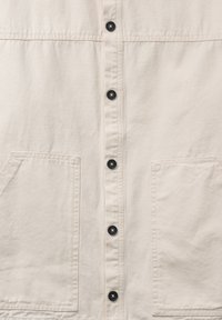 Devant d'une chemise en coton beige avec six boutons noirs alignés sur le centre et deux grandes poches carrées plaquées en bas.