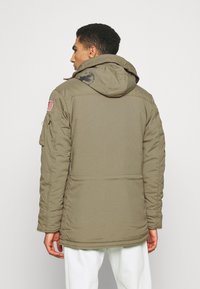 Giacca puffer khaki con cappuccio, caratterizzata da una patch con la bandiera americana sulla manica e un logo sul retro. Texture morbida, vestibilità adattata.