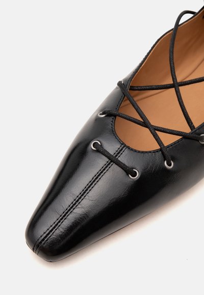 Chaussure plate en cuir noir à bout pointu avec des lacets noirs croisés passant à travers des œillets argentés et une doublure intérieure beige.