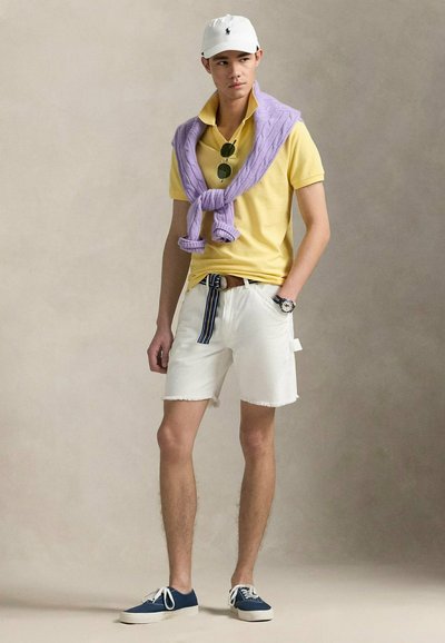 Polo giallo, maglione viola drappeggiato, pantaloni corti bianchi sfilacciati, scarpe da ginnastica blu e un cappello bianco. Gli accessori includono occhiali da sole e un orologio.
