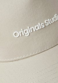 Bežinė kepurė su tekstūruota medžiaga, priekyje yra balti užrašai "Originals Studio". Matoma siuvimo linija aplink kraštą.
