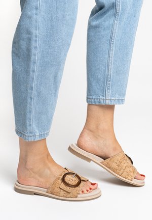 Piedi con jeans azzurri chiari e sandali beige a ciabatta con dettaglio fibbia rotonda, un piede leggermente sollevato su sfondo bianco.