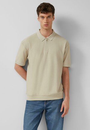 Jeune homme aux cheveux bruns bouclés portant un polo en maille beige à manches courtes et un jean bleu, debout devant un fond clair uni.