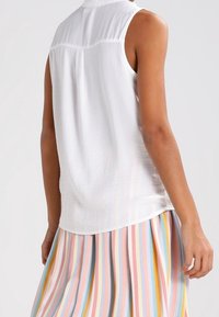 Femme portant un haut sans manches blanc et une jupe plissée à rayures verticales pastel en bleu, rose, jaune et orange, se tenant devant un fond uni.