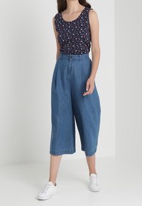 Ärmelloses, marineblaues Oberteil mit rot-weißen Mustern, kombiniert mit hellblauen, weit geschnittenen Jeans-Culottes und weißen Sneakers.