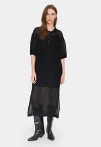 Rochie tricotată neagră cu mânici scurte și guler. Prezintă un tiv inferior transparent și patru nasturi metalici la nivelul pieptului.