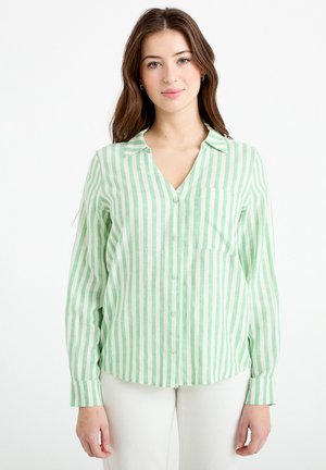 Młoda kobieta z długimi brązowymi włosami, ubrana w koszulę z pionowymi zielono-białymi paskami na guziki oraz białe spodnie, stoi na tle białego tła.