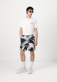 T-shirt bianca a maniche corte con logo, pantaloni corti neri con stampa tropicale blu e crema, sneakers bianche e occhiali da sole.