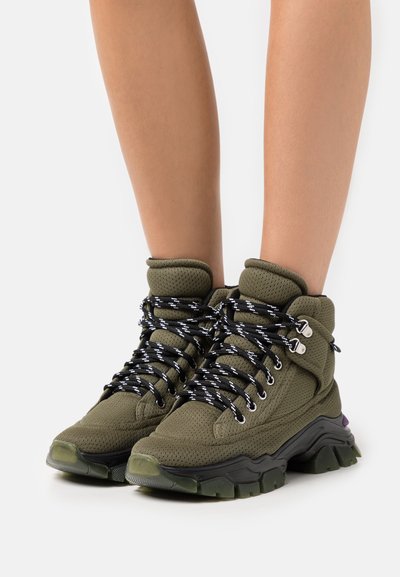 As sapatilhas altas verde-azeitona apresentam um design em malha, atacadores em padrão preto e branco, e uma sola preta grossa com um detalhe roxo.