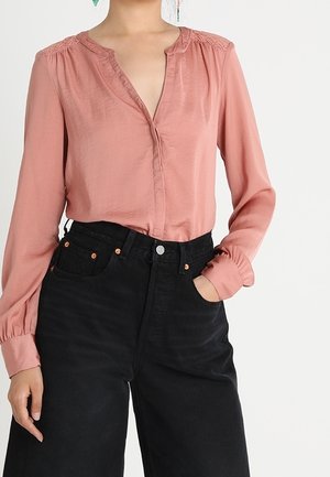 Woman wearing a dusty pink long-sleeve blouse tucked into high-waisted black wide-leg pants, hand in pocket.