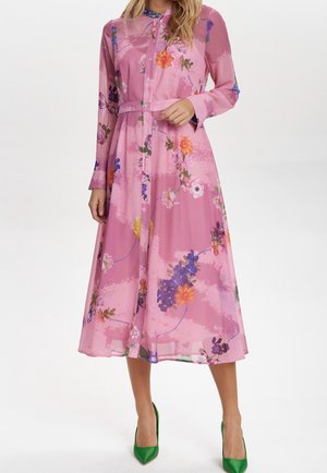 Femme portant une robe rose à manches longues à motif floral avec des boutons sur le devant et des talons hauts verts, debout devant un fond uni.