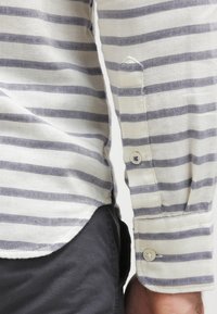 Camicia a righe in bianco sporco e navy, con polsini a bottone e una sottile trama. Il tessuto è leggero e dal design classico.