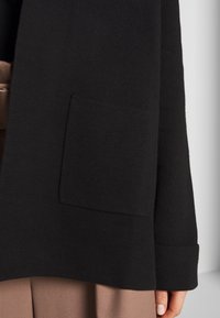 Svart stickad cardigan med bred krage, stora framfickor och ribbade ärmslut, lagd över ett ljusbrunt plagg.