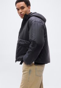 Νέος άνδρας που φοράει μαύρο φουσκωτό μπουφάν με κουκούλα και μπεζ παντελόνι, στέκεται με τα χέρια στις τσέπες, κοιτάζοντας την κάμερα.