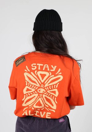 T-shirt en coton orange avec un grand graphisme crème au dos, présentant un design géométrique et le texte "STAY ALIVE." Bonnet noir porté.
