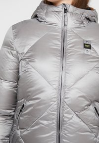 Grijze pufferjas met een ritsluiting, diamanten gestikt patroon, verstelbare capuchon en twee zijzakken met rits. Bevat een logo op de patch.