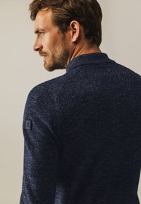 Marineblauw gebreide trui met een gestructureerde afwerking, voorzien van een polo kraag en een geborduurd logoembleem op de linker schouder.
