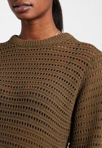 Olivgrüner Strickpullover mit offener Maschenstruktur, der horizontale Rippmuster sowie einen klassischen Rundhalsausschnitt aufweist. Sichtbare Details des strukturierten Fabrikmaterials.