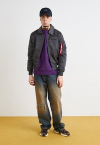 Mężczyzna noszący czapkę Nike w kolorze czarnym, czarną kurtkę bomber z czerwoną metką, fioletową bluzę, wyblakłe podarte dżinsy i czarne sneakersy na żółtej podłodze.
