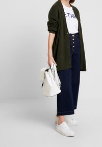 Green long cardigan, white graphic tee, high-waisted dark denim culottes, and white sneakers. White backpack with adjustable straps.