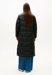 Schwarze Daunenjacke mit langem Schnitt, ausgestattet mit horizontalen Steppnähten und glänzender Textur. Kombiniert mit blauen weit geschnittenen Jeans.