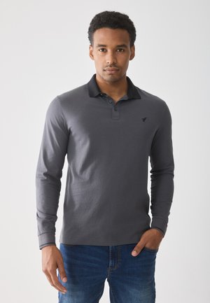 Camisa tipo polo de manga larga en algodón gris oscuro con un cuello negro, tapeta de tres botones y un pequeño logo en el pecho. Se lleva con vaqueros azules.