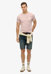 Camiseta de manga corta rosa claro con bolsillo, pantalones cortos de mezclilla azules, suéter de punto color crema anudado en la cintura, zapatillas deportivas blancas y calcetas con estampado.