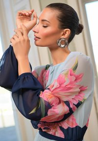 Fließendes Kleid mit transparenten, gepufften marineblauen Ärmeln und einem hellblauen Oberteil, das mit großen rosa Blumenmustern versehen ist. Enthält einen runden, durchsichtigen Ohrring.