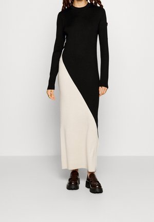 Frau trägt ein langes schwarz-weißes Kleid mit Farbblockmuster, langen Ärmeln und dunklen Plateau-Loafers, steht vor einem einfarbigen Hintergrund.