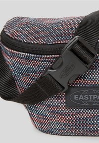 Ledvinka s texturovanou látkou v modrých, růžových a černých odstínech. Má černý popruh a přezku a oválnou nášivku s logem Eastpak.