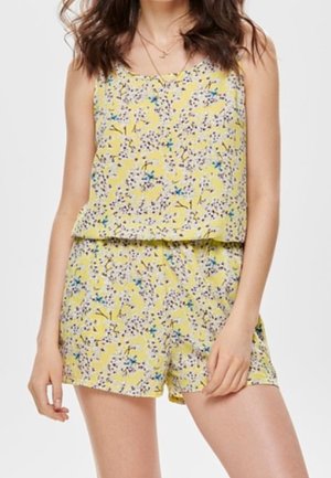 Kobieta ubrana w żółty, bezrękawnikowy kombinezon w kwiaty z niebieskimi i fioletowymi kwiatami, stojąca na jasnym, jednolitym tle.